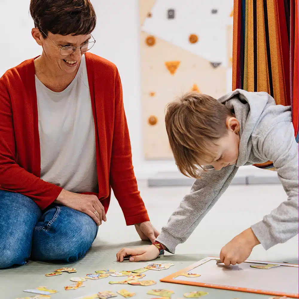 Lerntherapie Junge liegt in Bauchlage in der Hängematte und macht ein Puzzle. Daneben sitzt die Therapeutin Dorothe Schmidt.