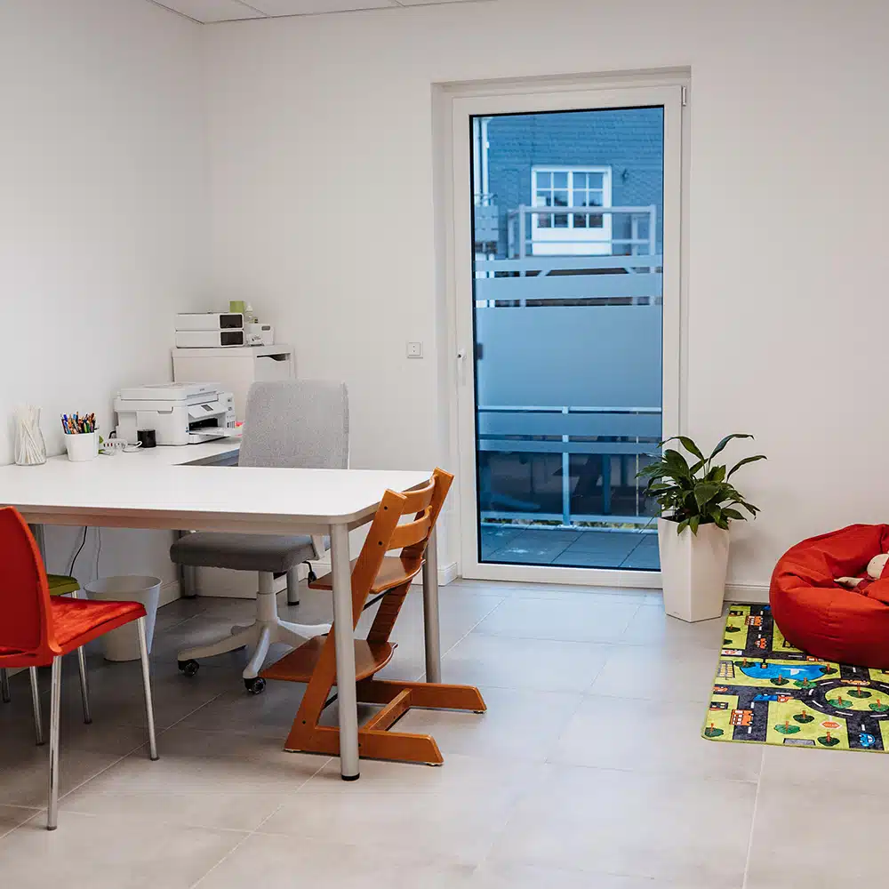 Kindersprache - Raum Blick auf einen L-förmigen Schreibtisch mit Drucker, Drehstuhl und Sitzmöglichkeiten für Patienten. Rechts Sitzsack auf einem Teppich, im Hintergrund großes Fenster