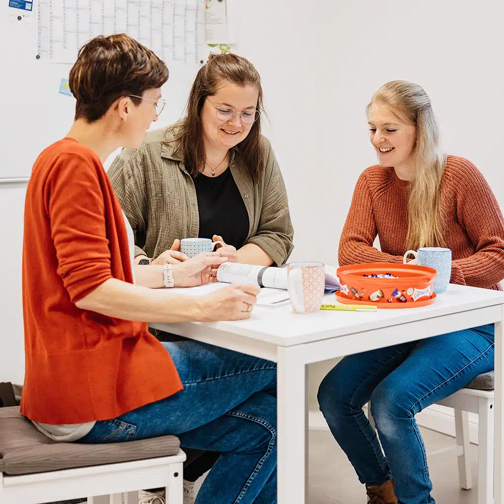 Carolin Eilinghoff, Dorothe Schmidt und Svenja Dommach sitzen während einer Besprechung mit Kaffee und Süßkeiten am Tisch im Personalraum.